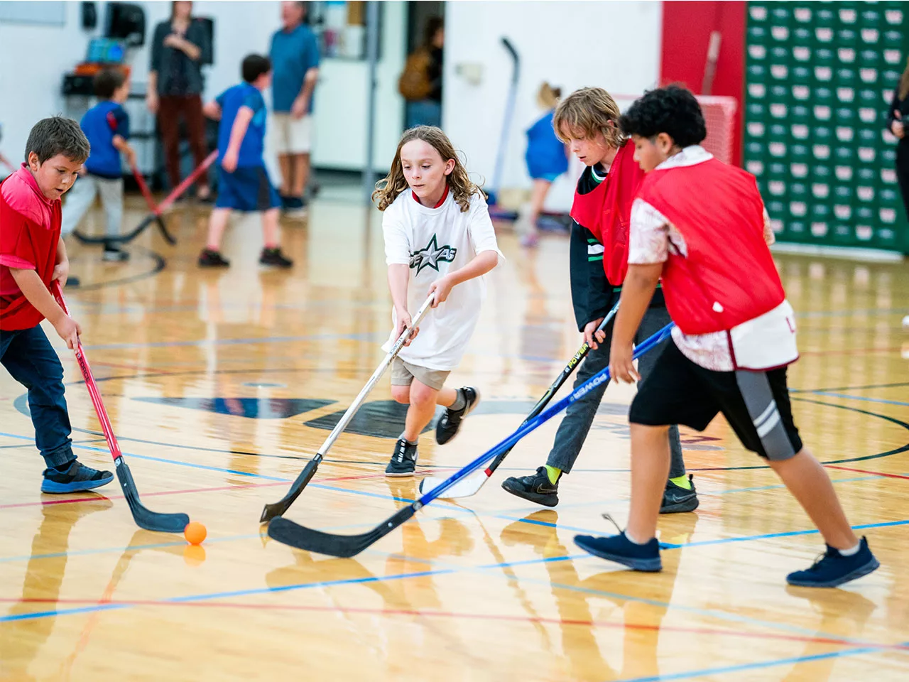 A group of children play a game of indoor hockey on a hardwood floor.