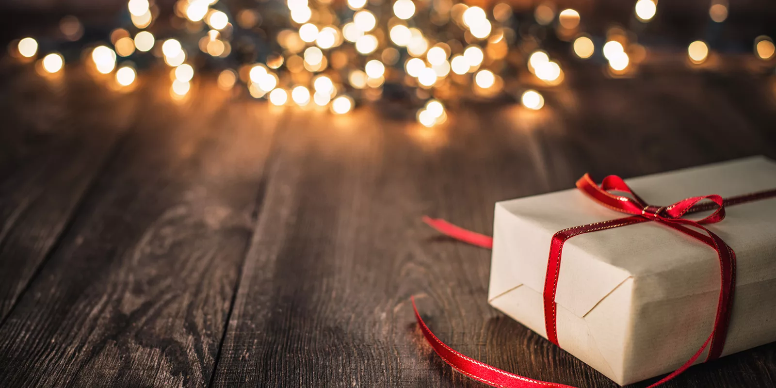 A wrapped gift is set on a wooden floor. String lights twinkle in soft focus in the background.