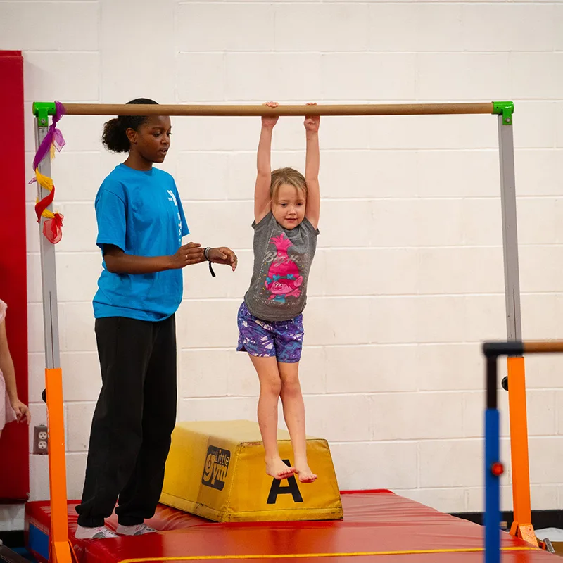 A child uses a gymnastics beam while a coach provides guidance.