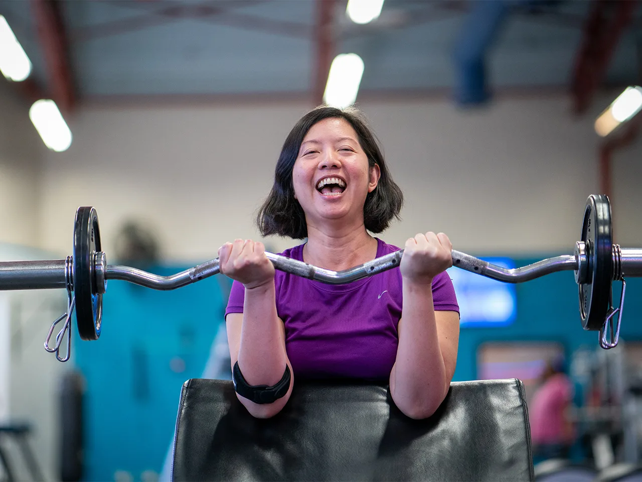 A woman smiles while flexing a barbell using a bicep machine.