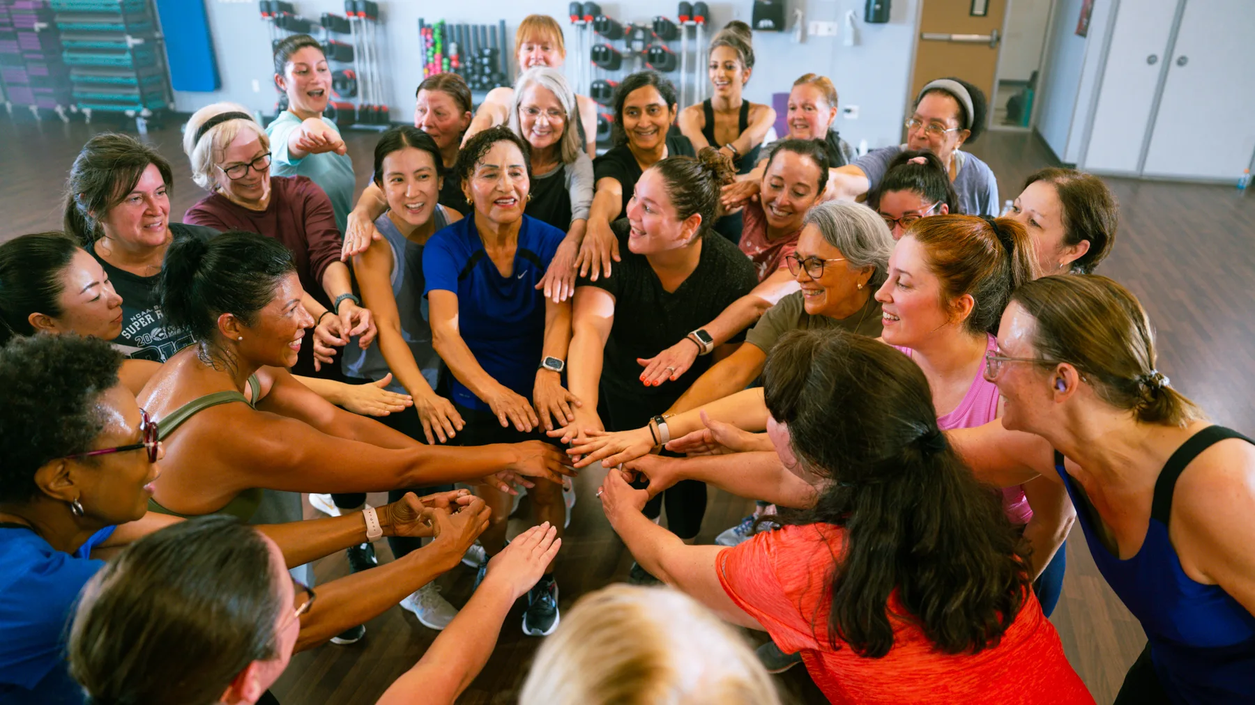 A smiling group of women come together with their hands in a circle after a group exercise class.