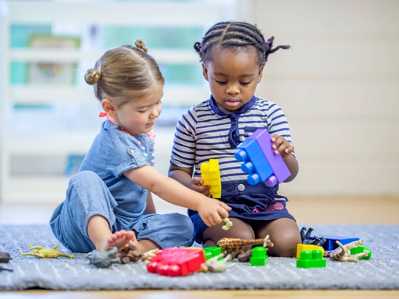 two toddlers kneel on a floor playing with toys