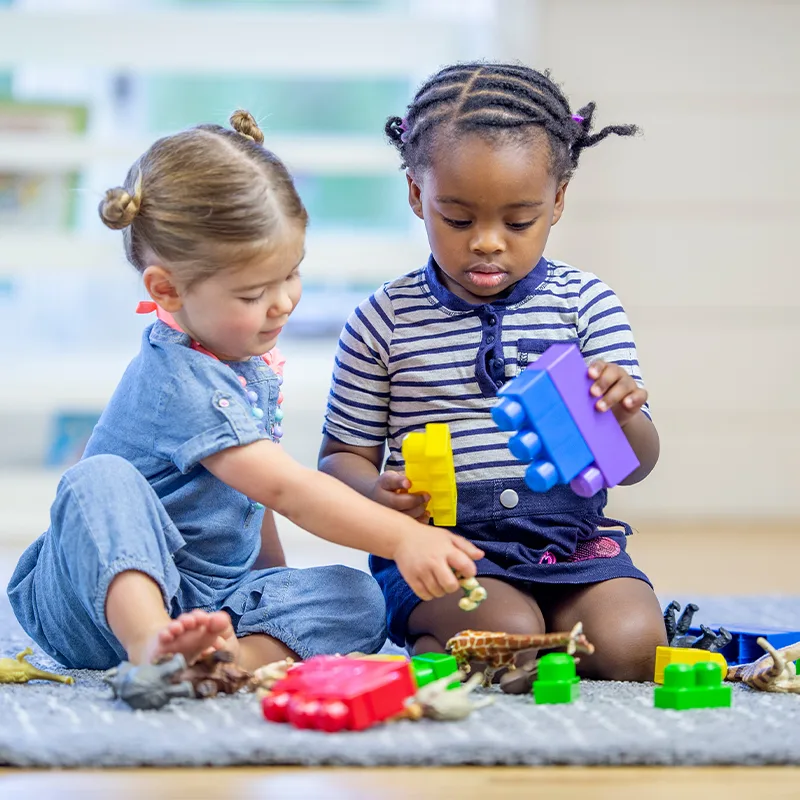 two toddlers kneel on a floor playing with toys