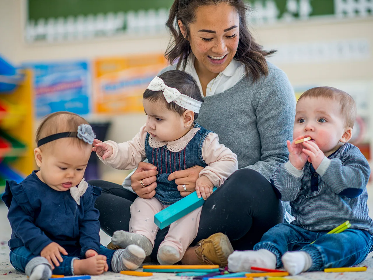 a teacher sits on a floor with a baby on her lap and two babies next to her.