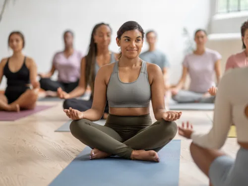 A woman sits cross-legged on a yoga mat in a group class.