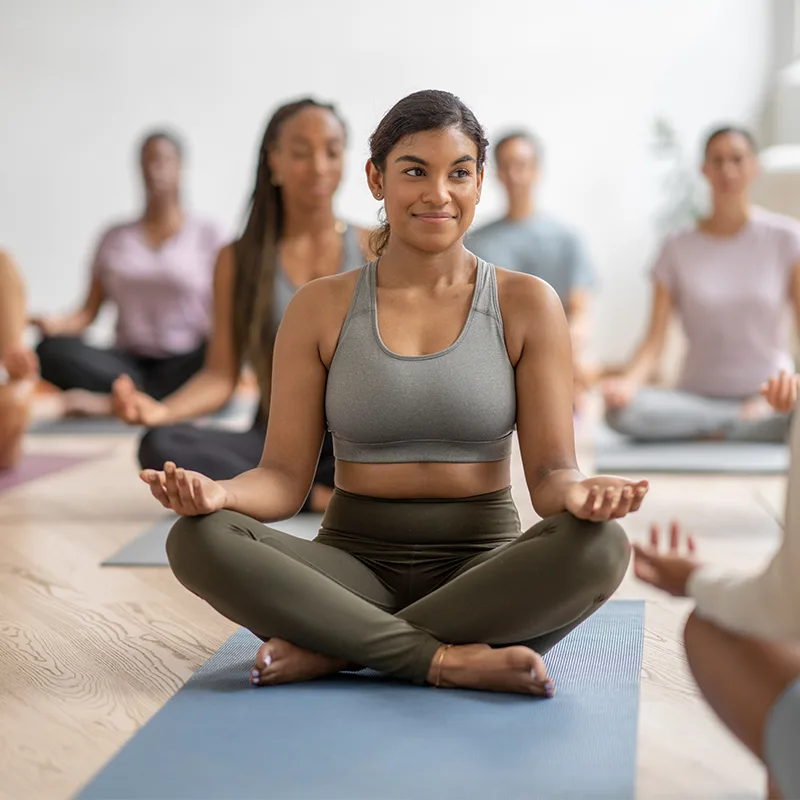 A woman sits cross-legged on a yoga mat in a group class.