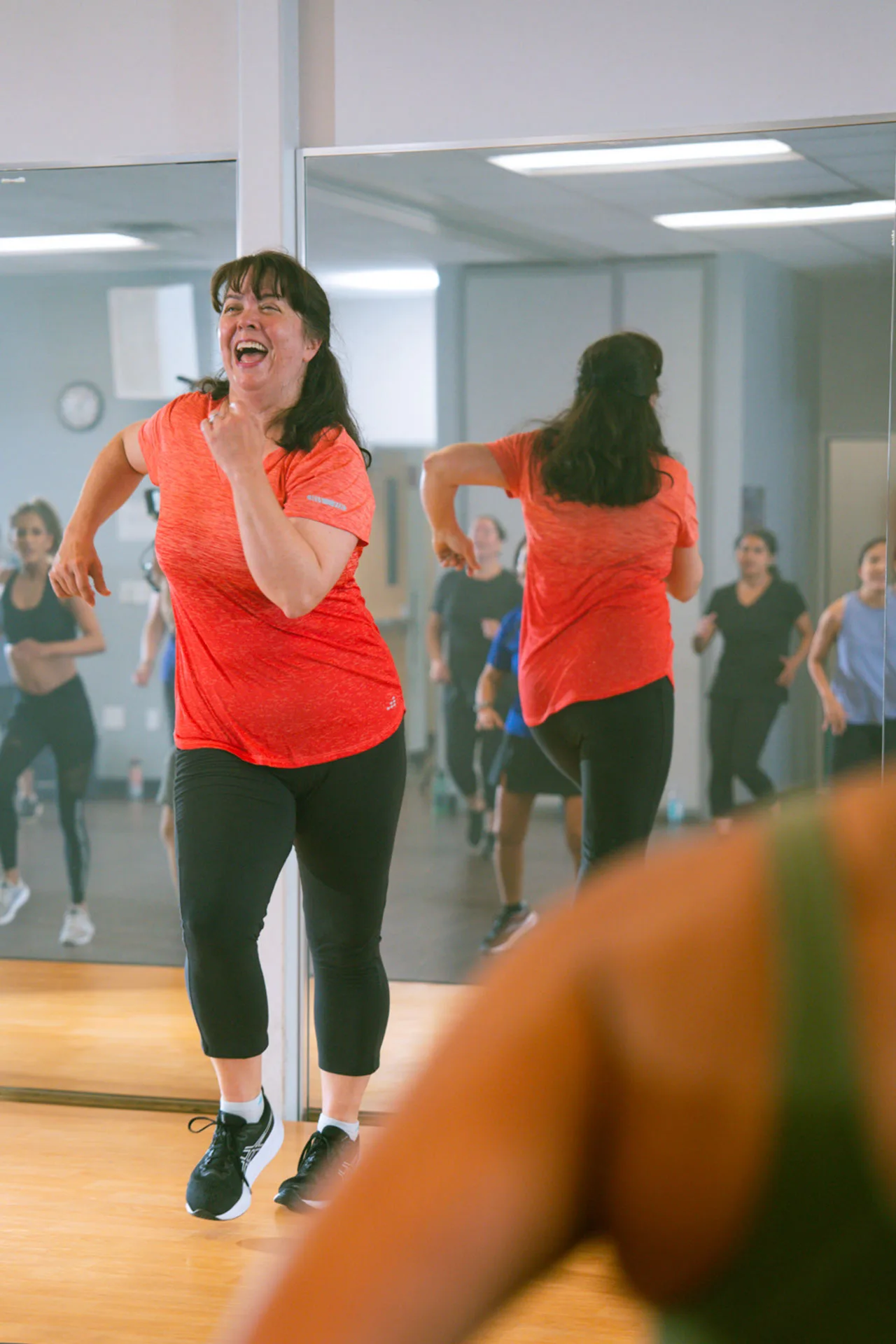 A woman smiles and dances with high energy as she leads a group fitness class.