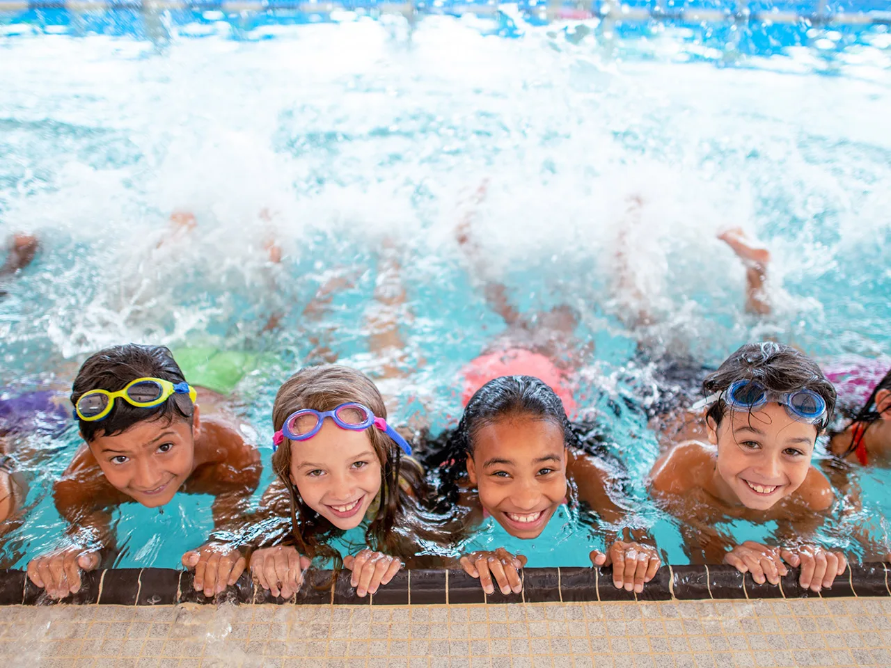 Children wearing goggles and colorful swimsuits cling to the edge of a pool and practice their kicking. The kids are smiling and having fun.