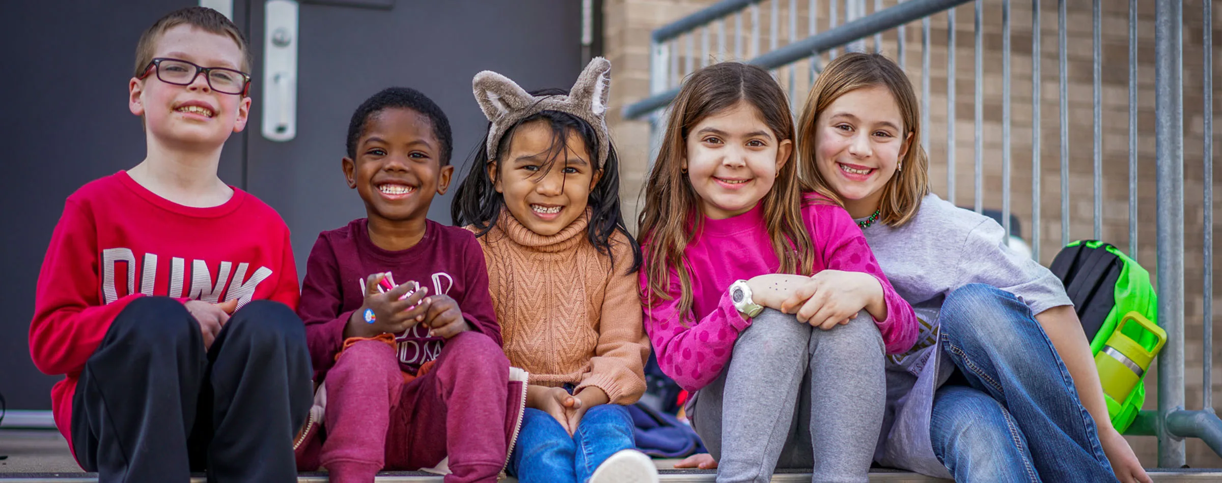 Five children sit together on the steps of their school smiling at the camera.