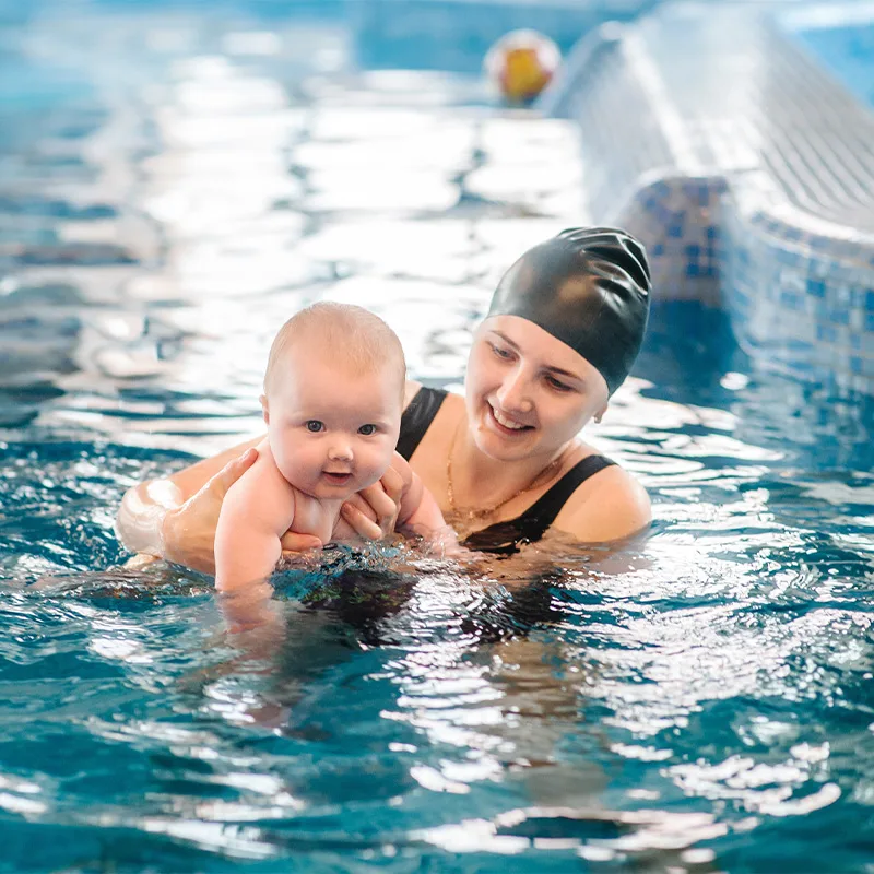 A mother and a baby participate in a swimming lesson in an indoor pool.