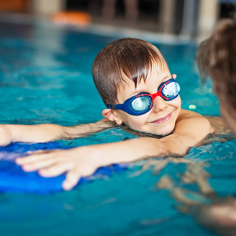 A young child wearing goggles participates in a swimming lesson in an indoor pool.