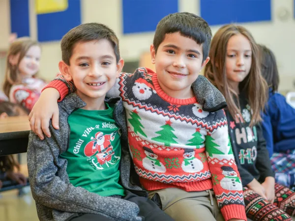 Two boys in holiday-themed clothing sit together smiling with their arms over each other's shoulders