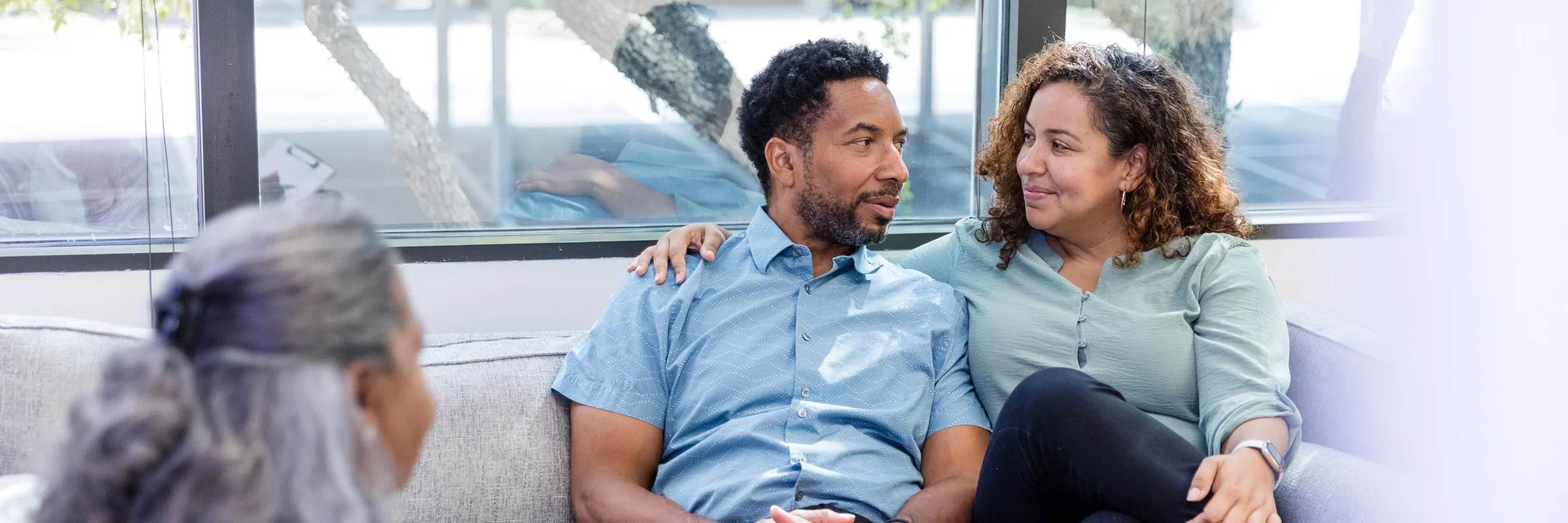 A couple sit on a sofa in a casual embrace, looking at one another.