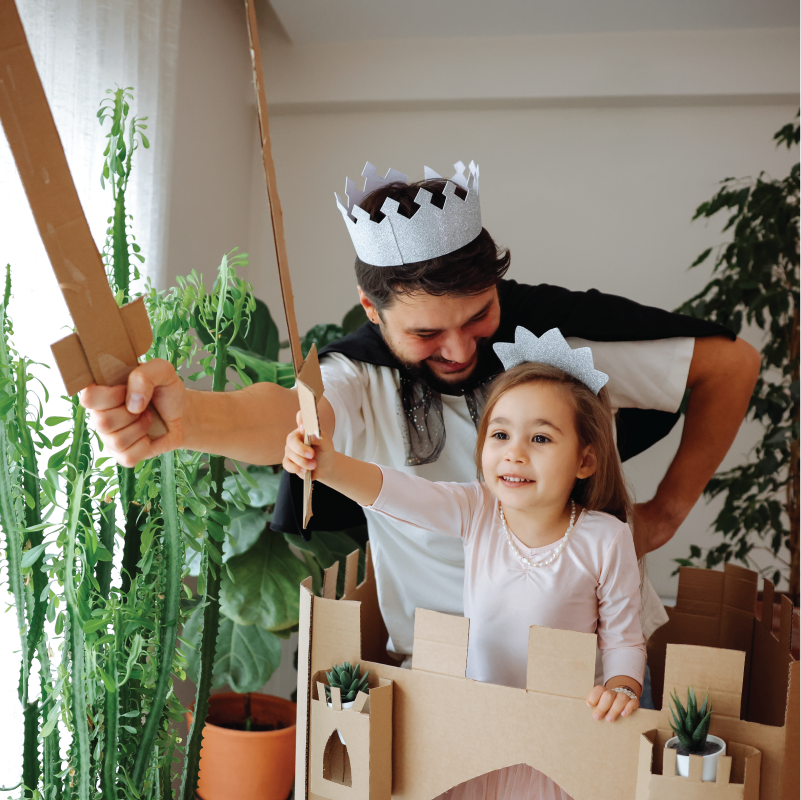 A father and his child dressed up in crowns and holding up cardboard swords pretend play