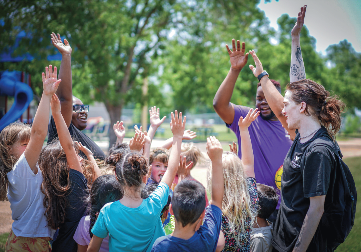 A group of staff and summer campers do a group cheer outside.