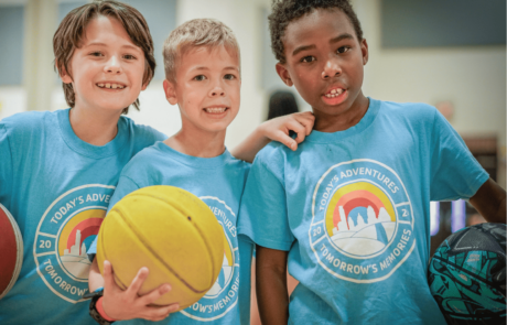 A group of three boys wearing blue summer camp t-shirts and holding basketballs look and smile at the camera.