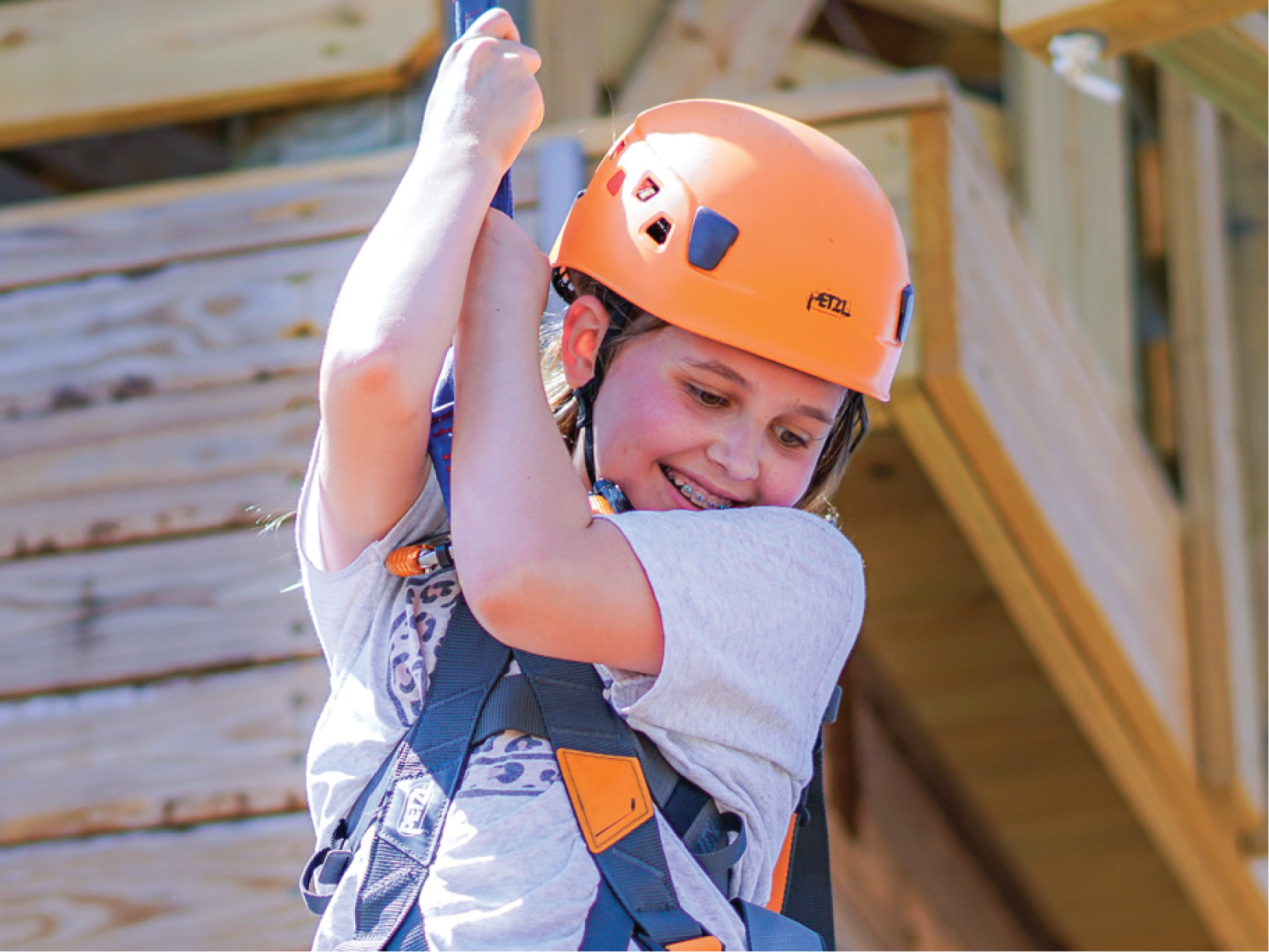 A child harnessed, wearing a helmet, and holding on to a rope smiles as she jumps down from a platform to the ground.