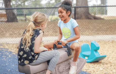 Two girls laugh and smile playing on a seesaw toy on the playground.