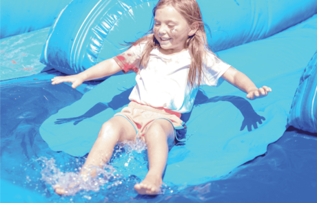 A girl smiles as she splashes at the end of an inflatable water slide on Olympic Day. She's wear a tie-dyed shirt and shorts.