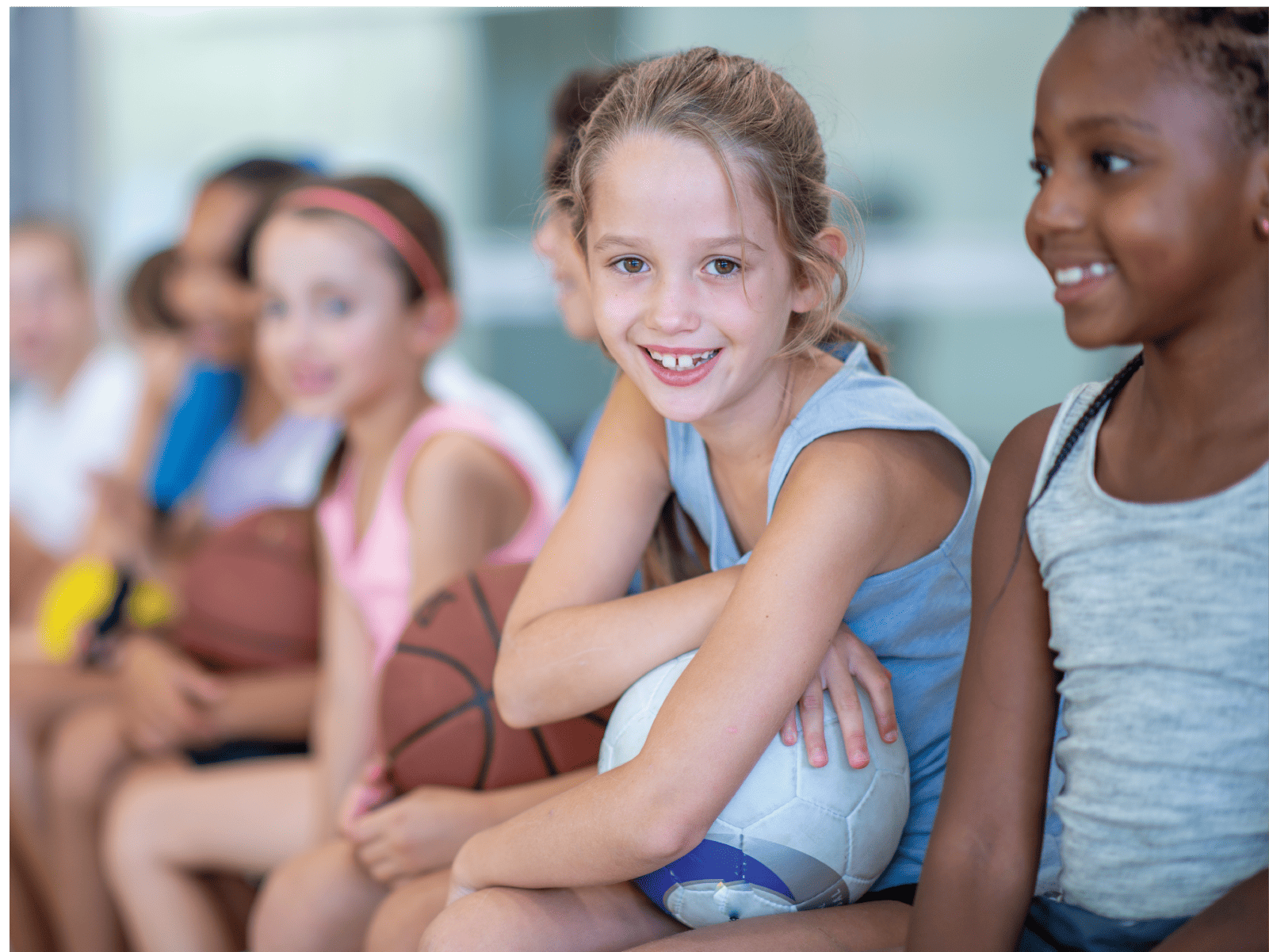 A group of children sit together holding basketballs waiting for instruction. One smiles at the camera.